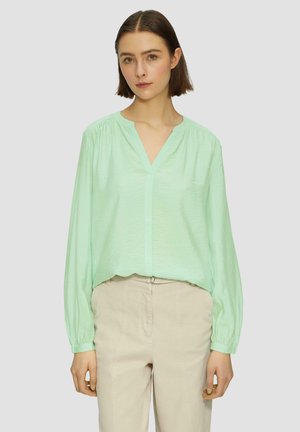 Muntgroene blouse met lange mouwen, een V-hals en geplooide schouders. Lichtgewicht, gestructureerde stof. Gecombineerd met beige broek.