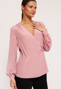 Różowa bluzka zakładana z satynowego materiału, z długimi bufiastymi rękawami i głębokim dekoltem w kształcie litery V. Guzik na rękawie dodaje charakterystycznego akcentu.