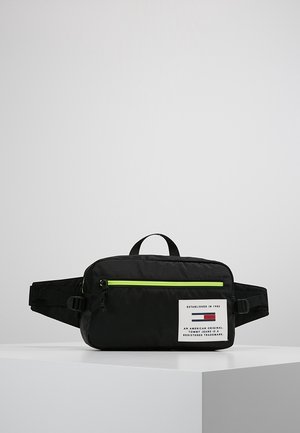 Czarna biodrówka z neonowozielonym zamkiem błyskawicznym, regulowanym paskiem, małym górnym uchwytem oraz białą naszywką z logo Tommy Jeans na przedniej kieszeni.