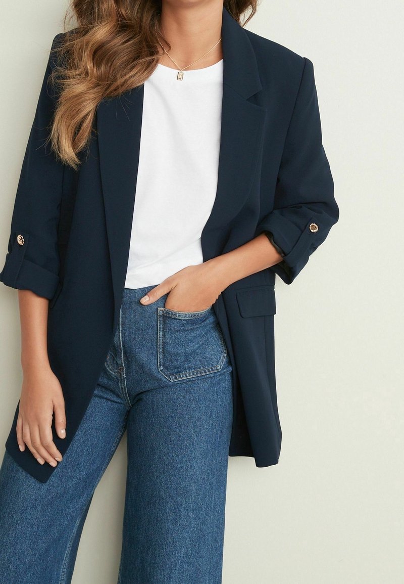 Blazer ajustado azul marino con mangas enrolladas, combinado con una camiseta blanca y unos vaqueros de mezclilla azul de tiro alto. Acentos de botones dorados en el blazer.