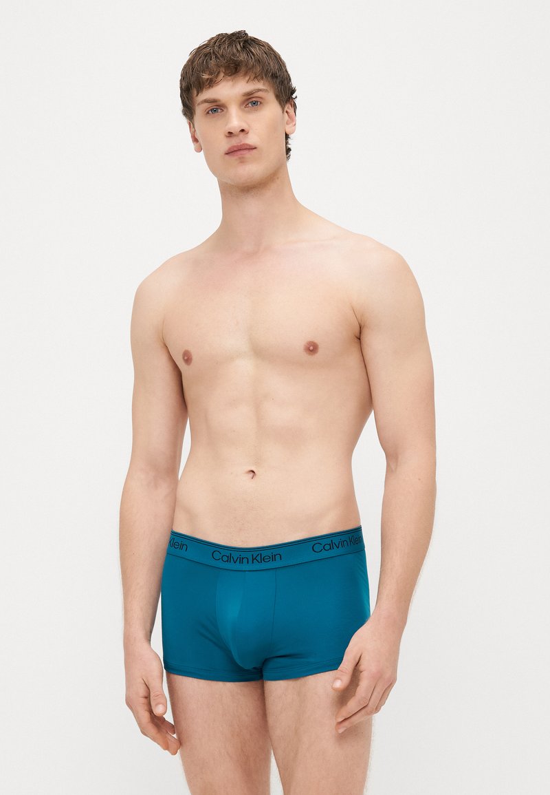 Tirkīzzils kokvilnas bokserbikses ar melnu jostu, uz kuras ir uzraksts „Calvin Klein”, tām ir ciešs pieguļošs un gluds audums. Modelis attēlā bez krekla.