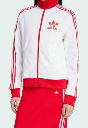 Persona vistiendo una chaqueta deportiva Adidas blanca y roja con rayas rojas en las mangas y logo rojo de Adidas, combinada con una falda roja con rayas blancas.