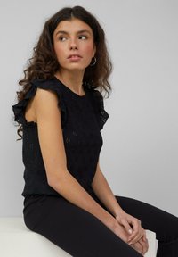 Zwarte top met ruche-accenten op de schouders, met oogjespatronen en een getailleerd ontwerp. Model droeg het met een zwarte broek.