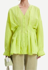 Camicia leggera a maniche lunghe di colore verde lime, con scollo a V, abbottonatura frontale e dettagli arricciati in vita. Tessuto texturizzato.