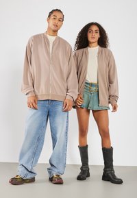 Light brown bomber jackets with zip closures, paired with a beige shirt, blue wide-leg jeans, and black knee-high boots with hardware accents.