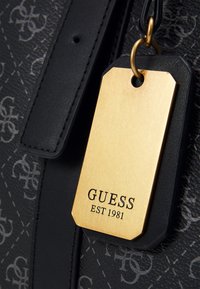 Borsa nera con superficie testurizzata e motivo a monogramma. Include un'etichetta dorata con inciso "GUESS" e "EST 1981".