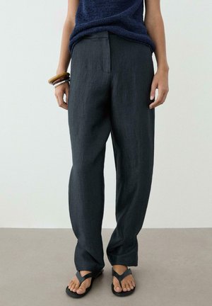 Person iført mørke løstsiddende bukser, navy ærmeløs top, sorte sandaler og flere armbånd på venstre håndled, står på grå gulv.
