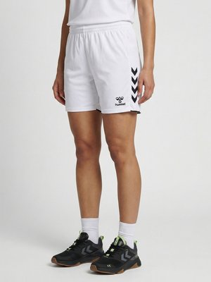 Person iført hvide hummel sportsshorts med sorte chevronstriber og sorte atletiske sko med hvide sokker, stående på gråt gulv.