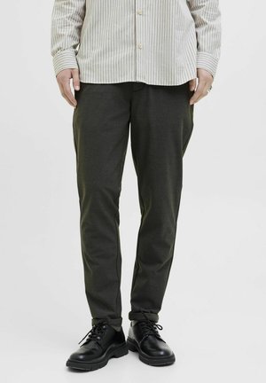 Dunkelgrüne Hose mit schmal zulaufendem Schnitt, aus einem glatten Material gefertigt. Kombiniert mit schwarzen Schnürschuhen, die eine strukturierte Sohle haben.