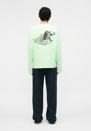 Άτομο με σγουρά μαλλιά φοράει ανοιχτό πράσινο μακρυμάνικο μπλουζάκι με μαύρο καρτούν χαρακτήρα που τρέχει και κείμενο, συνδυασμένο με χαλαρό σκούρο παντελόνι, στέκεται όρθιο.