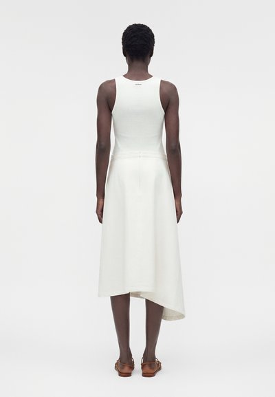 Γυναίκα με κοντά σγουρά μαλλιά που φοράει λευκό αμάνικο ριπ τοπ και ασύμμετρη λευκή φούστα, φαινομένη από πίσω, με καφέ σανδάλια.