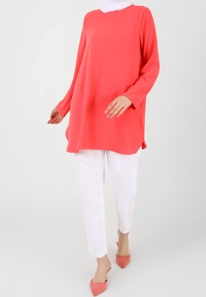 Femme portant une tunique corail à manches longues, un pantalon blanc, des talons corail à bout pointu et un foulard blanc sur la tête, debout devant un fond clair uni.