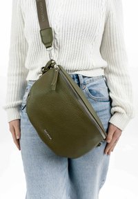 Olive green leather crossbody bag with pebbled texture, zip closure, and adjustable strap, featuring brand name embossed on the side.