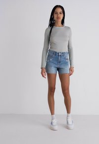 Maglietta grigia chiara a maniche lunghe abbinata a pantaloni corti in denim blu chiaro con bordi sfrangiati e ricami floreali. Il modello indossa scarpe da ginnastica bianche.