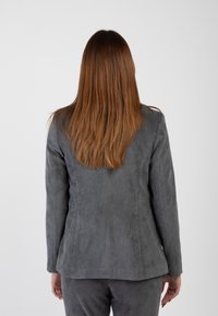 Donna con lunghi capelli castani, che indossa un blazer in corduroy grigio e pantaloni abbinati, in piedi di spalle su uno sfondo neutro.
