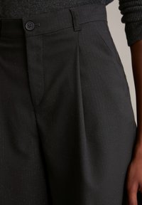 Zwarte op maat gemaakte broek van gestructureerde stof met subtiele streepjes. Voorzien van een knoopsluiting en twee zijzakken.