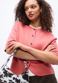 Fiatal nő göndör hajjal, rózsaszín gombos felsőben és fehér ingben, fekete-fehér mintás kézitáskát tartva, karját összefonva.