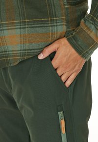 Rutiga skjortor i grönt med orange detaljer, tillsammans med mörkgröna byxor som har en zip-ficka och en slät yta.