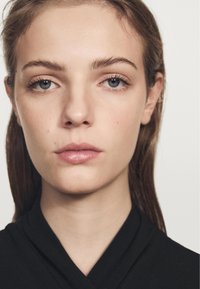 Detailní záběr na obličej osoby se světlou pletí, rovnými hnědými vlasy a neutrálním makeupem. Na sobě má černý texturovaný oděv.