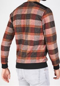Uomo che indossa un maglione a maniche lunghe con un motivo a quadri rosso, marrone, nero e rosa, abbinato a jeans grigi chiari, visto da dietro.