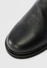 Svart læderstøvle med struktureret overflade, rund tå og en forhøjet søm langs sålen. Mat udseende med en glat kant.