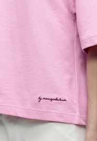 Ανοιχτό ροζ βαμβακερό πουκάμισο με κοντά μανίκια και σκούρο κεντημένο κείμενο "by marcopolobadrin" κοντά στο τελείωμα, συνδυασμένο με ανοιχτόχρωμο παντελόνι.