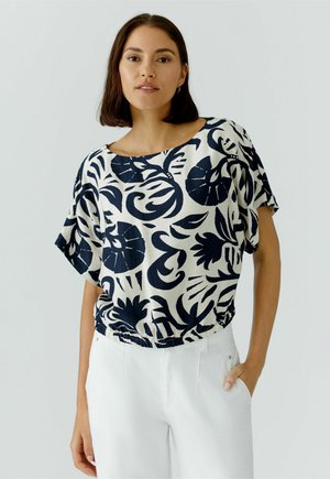 Jeune femme aux cheveux foncés mi-longs portant un chemisier blanc à grands motifs floraux bleu foncé et un pantalon blanc.