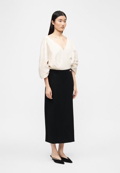 Γυναίκα με μακριά μαύρα μαλλιά, φοράει μια άνετη λευκή μπλούζα με V-λαιμόκοψη, μαύρη μίντι φούστα ψηλόμεση και μαύρα παπούτσια με μυτερή μύτη, όρθια.