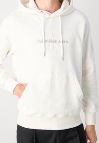 Person som bär en krämfärgad Calvin Klein Jeans-hoodie med framficka och dragsnören, handen i fickan, i kombination med svarta byxor.