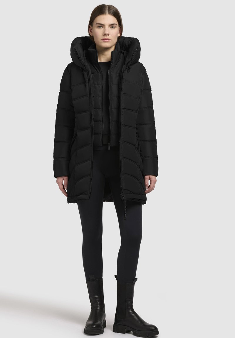 Svart pufferjacka med hög krage, flerpanelig design och dragkedjestängning. Bärs över svarta leggings och ankelstövlar.