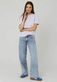 T-shirt violet clair avec un col rond, associé à un jean bleu taille haute à jambe large. Le mannequin porte des ballerines bleues.