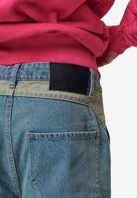 Denim bikses ar gaiši zilu mazgāšanu, ar melnu ādas uzlīmi aizmugures jostasvietā ar iepresētu logotipu un izliektiem kabatām. Redzama rozā sporta krekls.