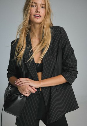 Blondynka w czarnej marynarce w cienkie prążki na czarnej bluzce, trzyma czarną skórzaną torebkę na szarym tle.