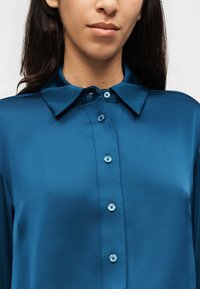 Κοντινό πλάνο ενός ατόμου που φοράει μία λαμπερή μπλε πουκαμίσα με κουμπιά και κλασικό γιακά, με εμφανές το λαιμό και το κάτω μέρος του προσώπου χωρίς τα μάτια.