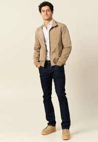 Beige leichte Jacke mit Reißverschluss und seitlichen Taschen, weißes gestreiftes Hemd, dunkelblaue Jeans und beige Wildlederschuhe. Glatte Textur, lässiges Design.