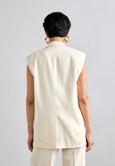Blazer sans manches crème avec un col haut, des épaules structurées et une coupe droite. Présente une couture centrale au dos et une texture subtile.