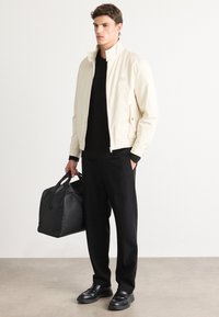 Man in cream jacket, black shirt and pants, black loafers holding black leather duffle bag, standing on light floor against white wall.