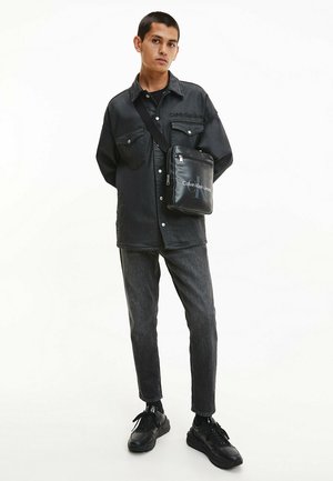 Schwarzes Jeanshemd mit zwei Brusttaschen, kombiniert mit dunkler Jeans und schwarzen Sneakers. Eine schwarze Umhängetasche zeigt das Calvin Klein-Logo.