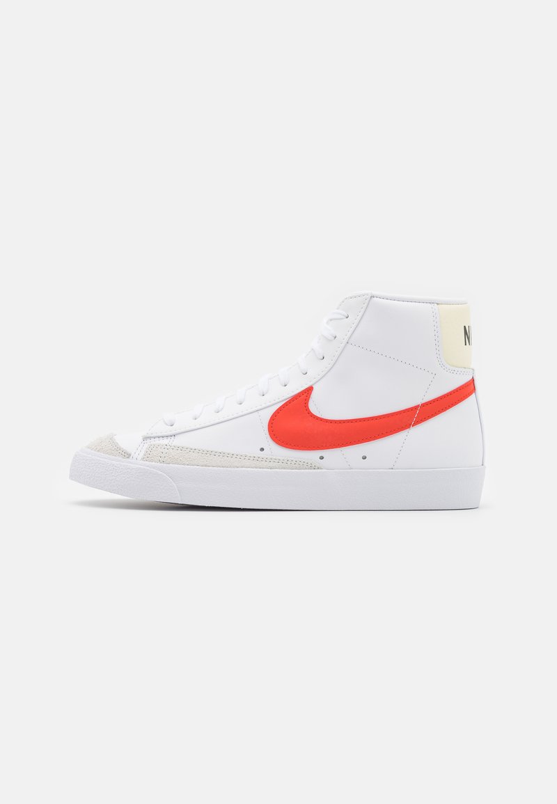 Bílé vysoké tenisky s červeným logem Nike, semišovým předním dílem a béžovým patním kloboučkem. Mají bílé tkaničky a texturovanou gumovou podrážku.