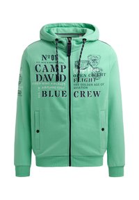 Sudadera con cremallera de color verde menta fabricada en material suave. Presenta gráficos de texto en negro, cordones y dos bolsillos frontales con cierres a presión.
