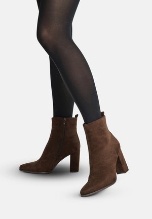 Ben iført gennemsigtige sorte strømpebukser og brune ruskindssko med blokhæle og sidelukning på en hvid baggrund.