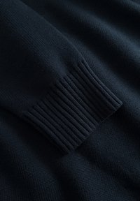 Détail d'un pull en maille bleu marine foncé montrant la texture des poignets côtelés et la finition lisse du tissu. Le tissage est serré avec un motif uniforme sur toute la surface.