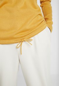 Žlutý pletený svetr s dlouhým rukávem a zaobleným lemem, kombinovaný s bílými kalhotami, které mají v pase řemínek. Hladká textura.