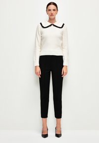 Pull en crème avec un col volant noir, associé à un pantalon noir ajusté et des escarpins noirs à bout pointu. Texture lisse et design ajusté.