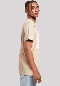 Camiseta de algodón beige de manga corta con texto blanco impreso, con cuello redondo y corte relajado, llevada con jeans de color azul claro.
