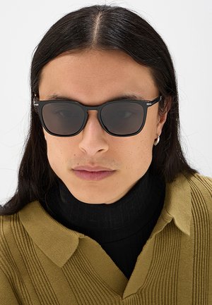 Persona con lunghi capelli neri che indossa occhiali da sole neri, orecchino a cerchio in argento, dolcevita nero e maglione a coste verde oliva.