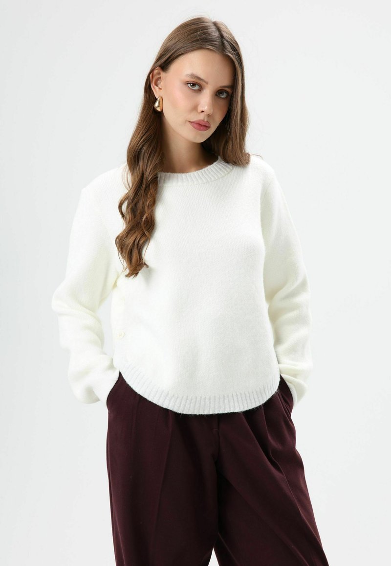 Pull blanc en maille avec encolure ronde, poignets et ourlet côtelés, associé à un pantalon bordeaux. Texture douce et design décontracté.