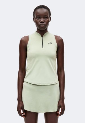 Donna che indossa un top sportivo verde chiaro senza maniche con zipper frontale e gonna abbinata, in piedi davanti a uno sfondo semplice.