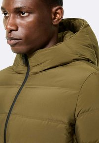 Giacca puffer verde oliva con cappuccio, dal tessuto liscio e una cerniera frontale con accenti neri. Il design è squadrato e moderno.