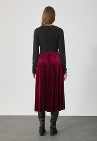 Top negru cu mânecă lungă și guler rotund, asortat cu o fustă de catifea burgundă, de lungime medie. Costumul are o talie strânsă și o textură netedă.